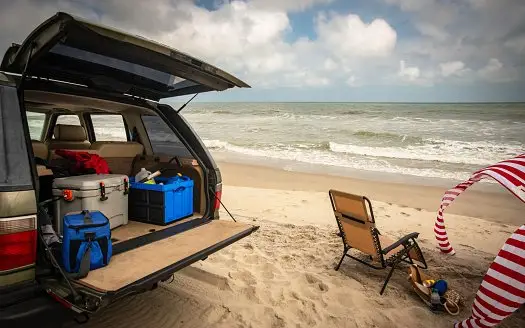 A beach with an open trunk of the vehicle and chairs